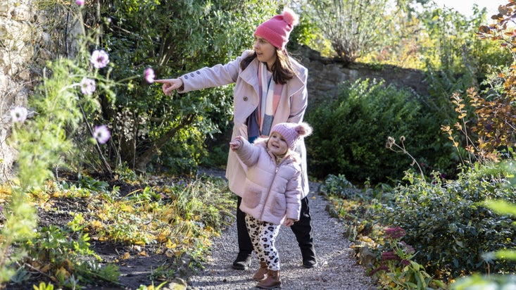 A woman and a small girl, both in pink woollen hats, point at flowers in a garden.
